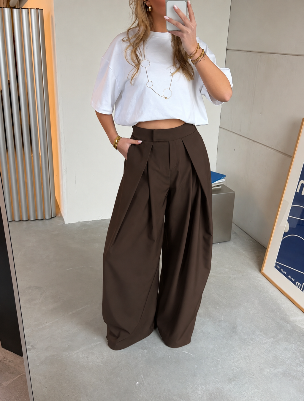 Dark brown wide-leg trousers with a high waist, deep front pleats, and an extra-wide flowing fit, styled with a white oversized T-shirt.